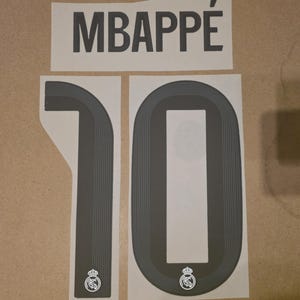 May include: A dark grey iron-on transfer set featuring the name MBAPPÉ in capital letters and the number 10. Each digit of the number 10 features a fine-line texture and the Real Madrid club crest logo at the base of the numeral.