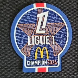 May include: A blue and white circular patch with the number "1" in the center. The patch has the text "Ligue 1" and "Champion 2024" in white. The McDonald's logo is in the center of the patch. The patch is surrounded by a pattern of stars and the text "Paris Saint Germain" repeated multiple times.