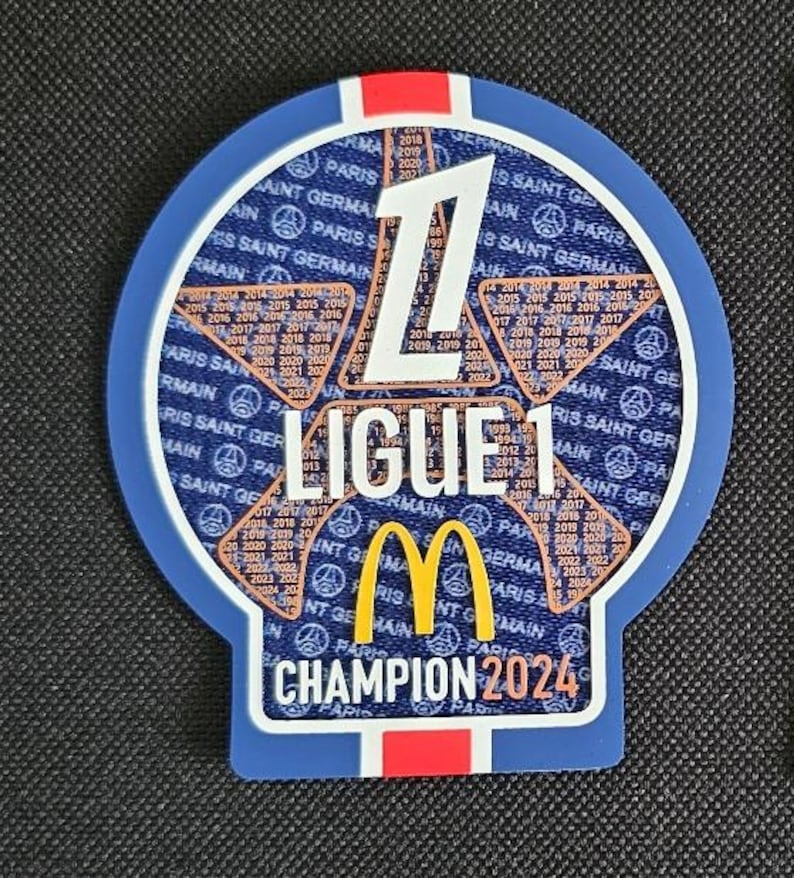 May include: A blue and white circular patch with the number "1" in the center. The patch has the text "Ligue 1" and "Champion 2024" in white. The McDonald's logo is in the center of the patch. The patch is surrounded by a pattern of stars and the text "Paris Saint Germain" repeated multiple times.
