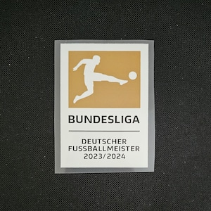 Op de afbeelding: Een witte patch met een gouden achtergrond waarop een silhouet van een voetballer te zien is die een bal trapt. De tekst "BUNDESLIGA" staat in het zwart onder de afbeelding. De tekst "DEUTSCHER FUSSBALLMEISTER 2023/2024" staat in het zwart onder de tekst "BUNDESLIGA".