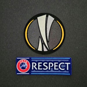 May include: A black and white embroidered patch with a stylised UEFA Champions League logo. The logo is surrounded by a yellow circle. Below the logo is a blue and white embroidered patch with the text "UEFA RESPECT".