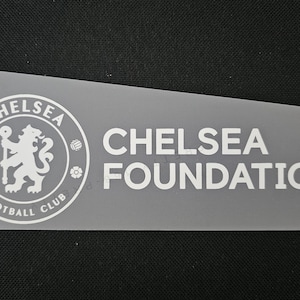 以下が含まれることがあります： 長方形の半透明ステッカー。チェルシーフットボールクラブのロゴと「CHELSEA FOUNDATION」の文字が白で表示されています。ロゴには、円形のエンブレムの中に杖を持ったライオンが含まれています。