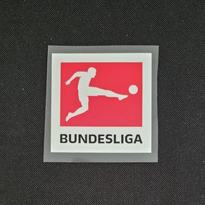 May include: A white silhouette of a soccer player kicking a ball on a red background with the text "BUNDESLIGA" in black below.