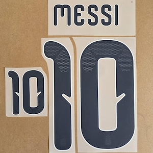 May include: A set of dark navy blue heat transfer decals featuring the name MESSI in a rounded font, along with two large number 10s. The numbers feature a white outline and a subtle dotted pattern at the top, with a small emblem at the base.