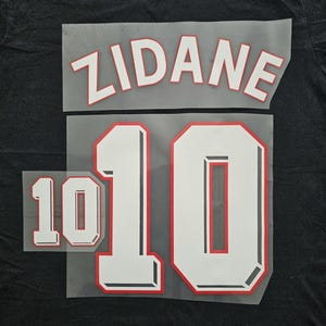 May include: A black t-shirt featuring a white and red heat transfer decal of the name ZIDANE in an arched font above the number 10. A smaller, separate white and red number 10 decal is positioned to the left of the larger number 10 graphic.