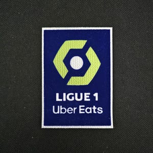 May include: A blue and white embroidered patch with a green hexagon logo and the text "LIGUE 1 Uber Eats".