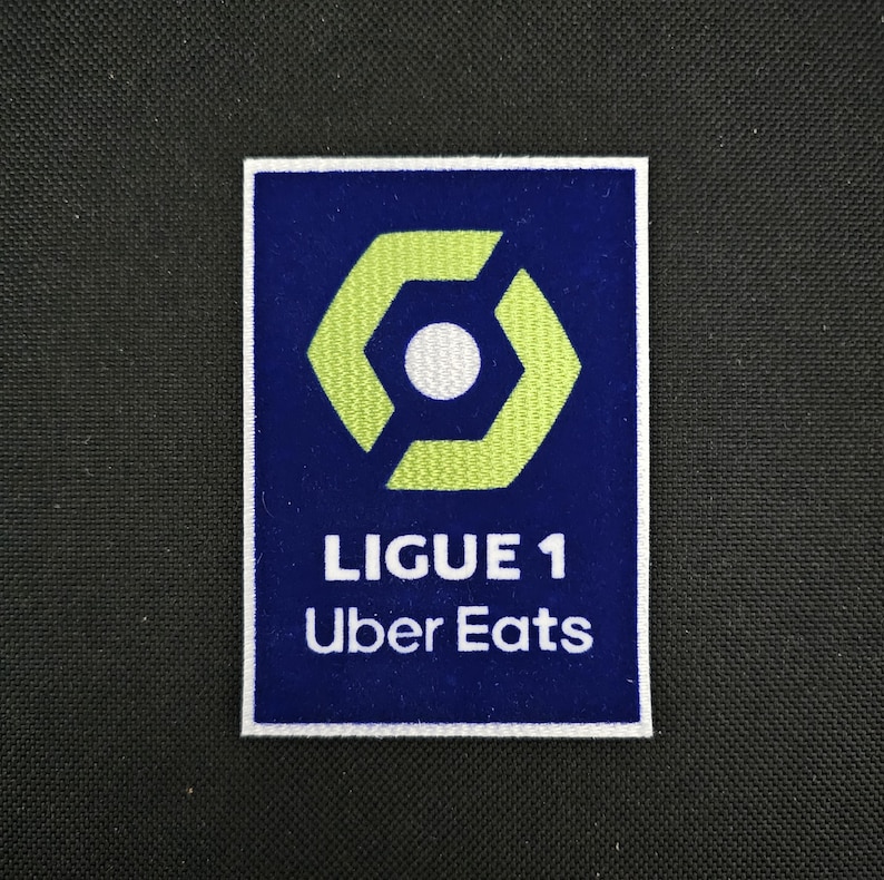 May include: A blue and white embroidered patch with a green hexagon logo and the text "LIGUE 1 Uber Eats".