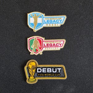 May include: Three embroidered patches featuring FIFA World Cup themes. Two patches display a gold silhouette of a player with the text FIFA WORLD CUP LEGACY, one for Messi in blue and white, one for Ronaldo in red and green. One patch shows the trophy with DEBUT FIFA WORLD CUP.
