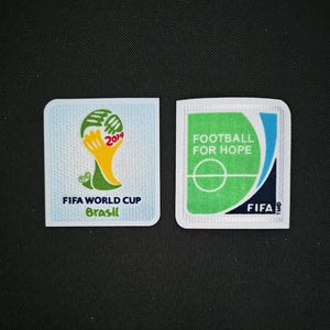 Può includere: Due toppe ricamate bianche. La toppa a sinistra presenta un pallone da calcio verde e giallo con il testo "FIFA World Cup Brasil 2014". La toppa a destra presenta un campo da calcio verde con il testo "Football for Hope" e il logo FIFA.