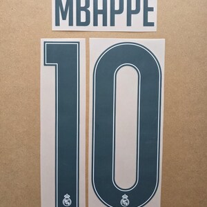 May include: A dark blue iron-on transfer set featuring the name MBAPPÉ in a tall, sans-serif font and the number 10. The number 10 includes a thin white outline and a small white Real Madrid club crest logo at the base of each digit.