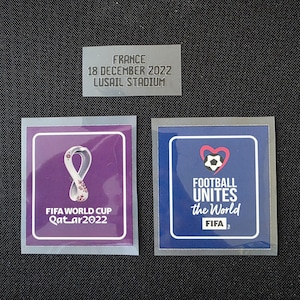 May include: Three square and rectangular stickers featuring FIFA World Cup Qatar 2022 branding. One sticker displays the purple FIFA World Cup Qatar 2022 logo, another shows the blue Football Unites the World logo, and a grey label reads FRANCE 18 DECEMBER 2022 LUSAIL STADIUM.