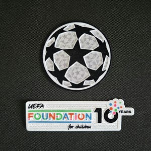 Könnte beinhalten: Ein schwarz-weißer Fußball-Patch mit Sternmuster. Darunter befindet sich ein weißer Patch mit dem Text "UEFA FOUNDATION for children 10 YEARS" in verschiedenen Farben. Die Patches befinden sich auf einem schwarzen Hintergrund.