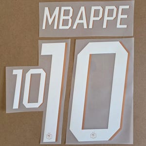 May include: A set of white iron-on heat transfer decals featuring the name MBAPPE in block letters and the number 10 in two different sizes. Each character has a thin copper-colored outline and a small hexagonal logo at the base of the numbers.