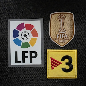 May include: Three embroidered patches including a rectangular LFP logo with a colourful circular emblem, a shield-shaped gold FIFA World Champions 2009 patch with a trophy graphic, and a square yellow patch featuring a red triangle and the number 3.