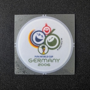 Può includere: Un adesivo circolare con il logo della Coppa del Mondo FIFA Germania 2006. Il design include tre volti sorridenti stilizzati in blu, rosso e verde, con il testo FIFA WORLD CUP GERMANY 2006 e un piccolo marchio 2002 FIFA TM.