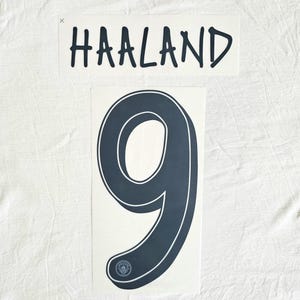 May include: A white sheet with two printed decals. The top decal reads "HAALAND" in black, stylized font. Below is a large number "9" in navy blue with a white outline and a small emblem at the bottom.