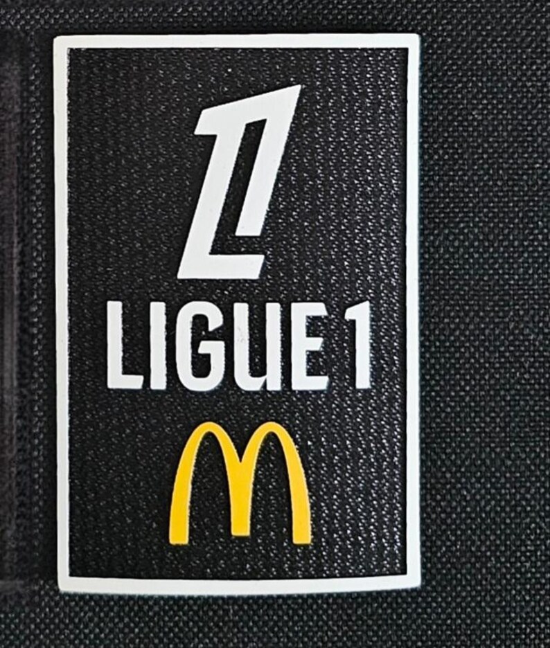 May include: A black and white patch with the text "Ligue 1" and a white number "1" above the text. A yellow McDonald's "M" logo is below the text.