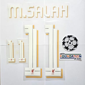 May include: A set of iron-on patches for a soccer jersey. The patches include the name "M. SALAH", the number "11", a soccer ball with the number "6", and a UEFA Foundation logo. The patches are white with gold outlines.