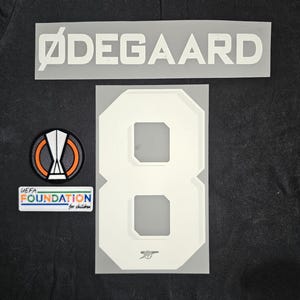 May include: White "ØDEGAARD" nameplate and number "8" with a white Arsenal crest on a black background. A small UEFA Foundation for Children logo is also visible.
