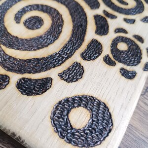 May include: Close-up of a wooden cutting board with a decorative, burnt design. The design features a spiral, circular, and crescent shapes in dark brown against the light wood grain. The board has a natural, rustic aesthetic.