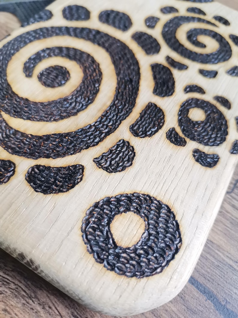 May include: Close-up of a wooden cutting board with a decorative, burnt design. The design features a spiral, circular, and crescent shapes in dark brown against the light wood grain. The board has a natural, rustic aesthetic.