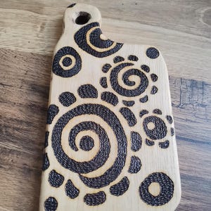 May include: A light-colored wooden cutting board with a handle and a series of dark, spiral and circular designs. The board has a smooth surface and a hole in the handle for hanging. The decorative design is burned into the wood.