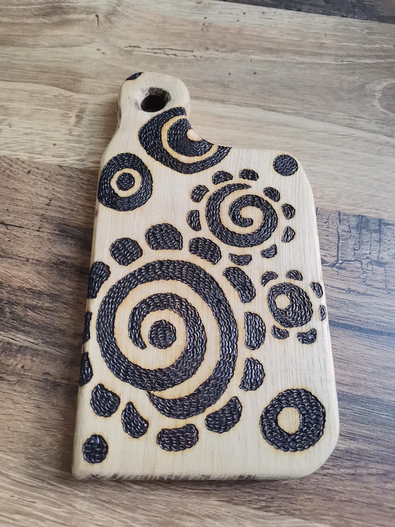 May include: A light-colored wooden cutting board with a handle and a series of dark, spiral and circular designs. The board has a smooth surface and a hole in the handle for hanging. The decorative design is burned into the wood.