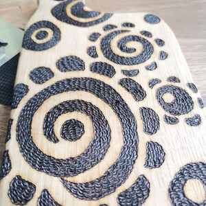 May include: A light-colored wooden cutting board with a handle. The board features a decorative pattern of dark, textured spirals and circular shapes. The design is burned into the wood, creating a contrast between the light wood and the dark, detailed patterns.