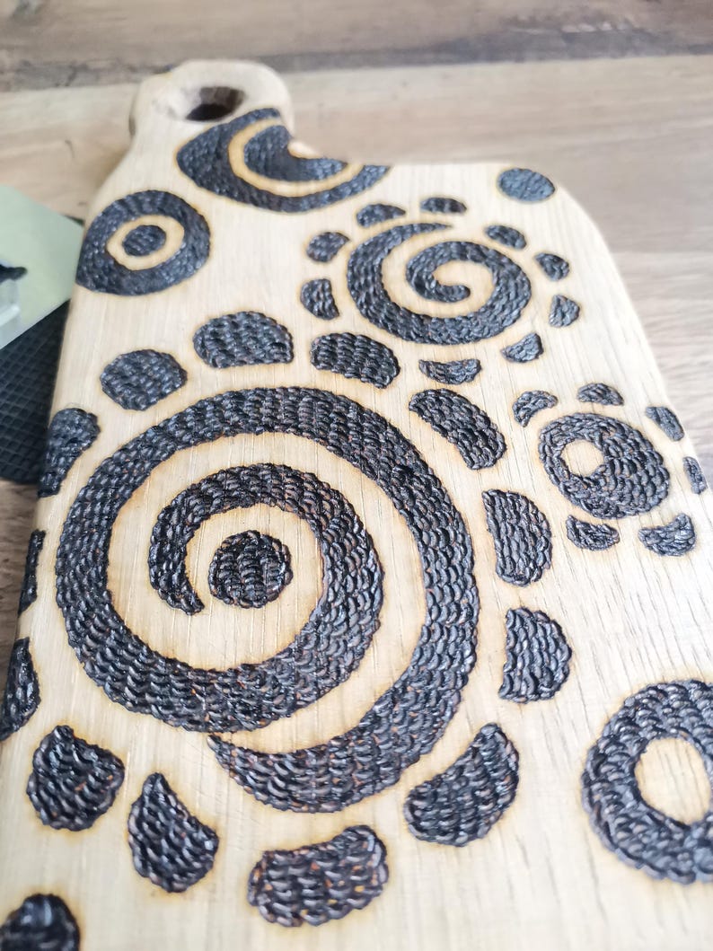 May include: A light-colored wooden cutting board with a handle. The board features a decorative pattern of dark, textured spirals and circular shapes. The design is burned into the wood, creating a contrast between the light wood and the dark, detailed patterns.