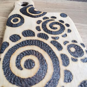 May include: Wooden cutting board with a handle and decorative black spiral and dot patterns. The board has a light wood color with dark, textured designs burned into the surface. The handle features a hole for hanging.