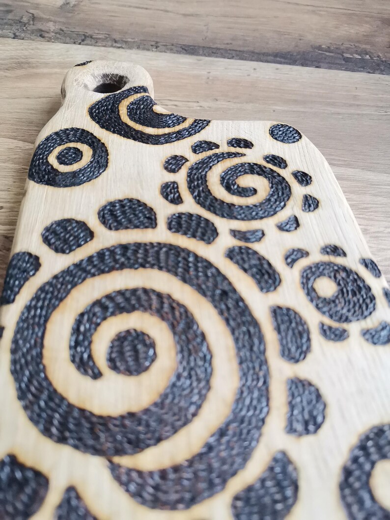 May include: Wooden cutting board with a handle and decorative black spiral and dot patterns. The board has a light wood color with dark, textured designs burned into the surface. The handle features a hole for hanging.