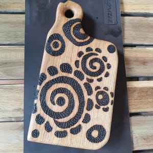 May include: Wooden cutting board with a handle, featuring a light brown surface and dark, spiraling, and circular patterns. The board has a unique shape and is suitable for kitchen use.