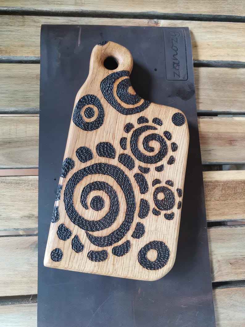 May include: Wooden cutting board with a handle, featuring a light brown surface and dark, spiraling, and circular patterns. The board has a unique shape and is suitable for kitchen use.