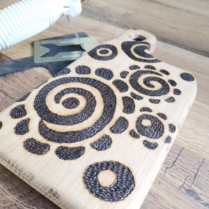 May include: A light-colored wooden cutting board with a handle, featuring a decorative pattern of dark brown spirals and circles. A wood-burning tool is in the upper left corner. The board is on a wooden surface.