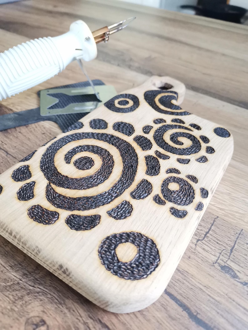 May include: A light-colored wooden cutting board with a handle, featuring a decorative pattern of dark brown spirals and circles. A wood-burning tool is in the upper left corner. The board is on a wooden surface.