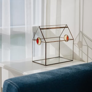 Może przedstawiać: Szklany terrarium w kształcie domu z metalową ramą i akcentami ze szkła witrażowego. Terrarium ma dwa małe, okrągłe okna ze szkła witrażowego z czerwonymi i żółtymi akcentami. Terrarium stoi na białym stole.