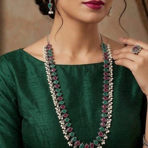 May include: A silver-toned necklace, earrings, and ring set. The jewelry features alternating green and red teardrop-shaped stones, accented with small white pearls. The woman is wearing a dark green top.