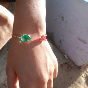 Può includere: Un braccialetto rigido in oro con una pietra preziosa verde e rossa. Il braccialetto è al polso di una persona.