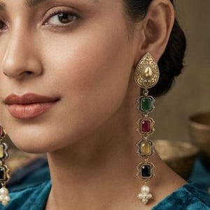 May include: Long, ornate earrings with a teardrop-shaped top and a series of colorful square gemstones. The gemstones are green, red, yellow, and black, with a pearl cluster at the bottom. The earrings are gold-toned.