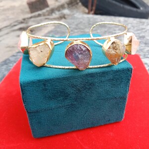 May include: Gold bangle bracelet with four different colored gemstones. The gemstones are set in a gold wire frame. The bracelet is on a green velvet box.