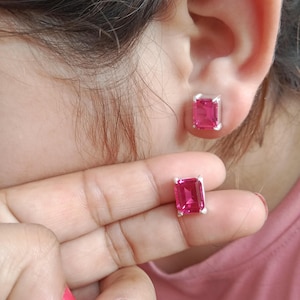 May include: A pair of stud earrings featuring rectangular, emerald-cut pink stones set in silver-toned metal prongs. One earring is worn in the earlobe, while the other is held between two fingers to display the vibrant pink gemstone clearly.