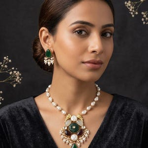 May include: A necklace featuring a strand of white baroque pearls with a large central pendant containing a green carved stone, dark red stones, and white pearls. Matching teardrop-shaped earrings feature a green stone and white pearl dangles.