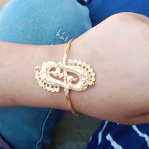 May include: A gold-colored bracelet with an ornate peacock design. The bracelet features a detailed, openwork peacock centerpiece and a delicate, twisted band. The design is symmetrical and the bracelet is worn on a wrist.