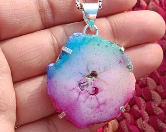 Peut inclure: Pendentif en agate multicolore avec une chaîne en argent. Le pendentif présente un mélange vibrant de teintes bleues, vertes, roses et violettes. La pierre est de forme irrégulière et a une surface polie, révélant ses motifs naturels.