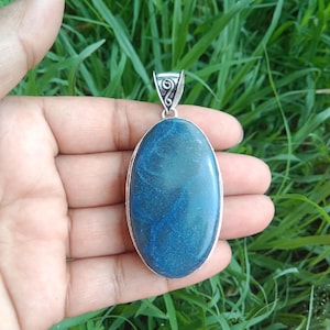 May include: An oval-shaped blue gemstone pendant set in a silver frame with a decorative metal bail. The stone features a smooth, polished surface with deep blue tones and subtle swirling patterns throughout the center of the cabochon.