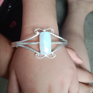 May include: A silver-toned cuff bracelet featuring a central, rectangular, milky white stone. The metal band splits into two thin strands on each side of the stone, accented with decorative, symmetrical scrollwork at the top and bottom edges.