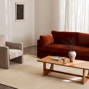 Puede incluir: Un sof&aacute; color &oacute;xido, un sill&oacute;n beige y una mesa de centro de madera clara. Sobre la mesa hay un jarr&oacute;n de cer&aacute;mica blanca con ramas secas oscuras, un cuenco beige peque&ntilde;o y un libro. Una alfombra beige grande cubre el suelo de la estancia.