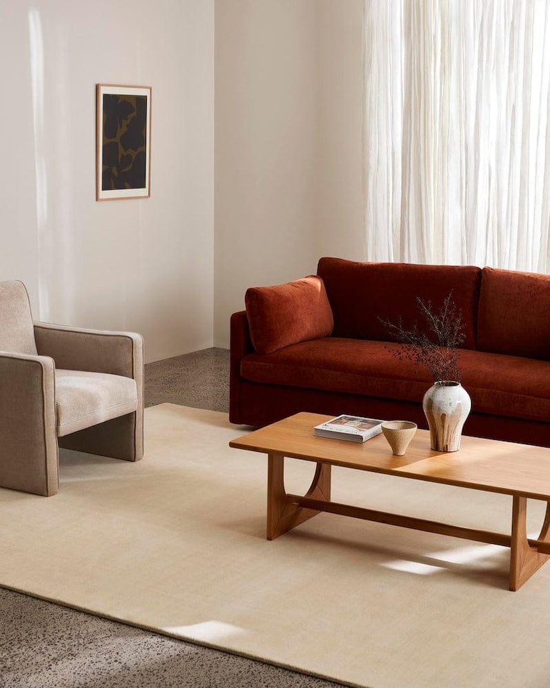 Puede incluir: Un sof&aacute; color &oacute;xido, un sill&oacute;n beige y una mesa de centro de madera clara. Sobre la mesa hay un jarr&oacute;n de cer&aacute;mica blanca con ramas secas oscuras, un cuenco beige peque&ntilde;o y un libro. Una alfombra beige grande cubre el suelo de la estancia.