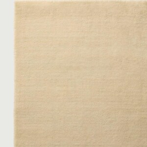 Puede incluir: Una alfombra rectangular de color beige claro s&oacute;lido. La superficie presenta una textura densa de pelo corto con una apariencia uniforme en toda la pieza. Los bordes son rectos y limpios, mostrando un tejido consistente en toda la alfombra.
