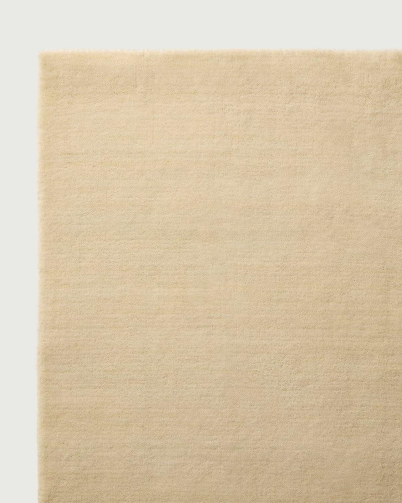 Puede incluir: Una alfombra rectangular de color beige claro s&oacute;lido. La superficie presenta una textura densa de pelo corto con una apariencia uniforme en toda la pieza. Los bordes son rectos y limpios, mostrando un tejido consistente en toda la alfombra.